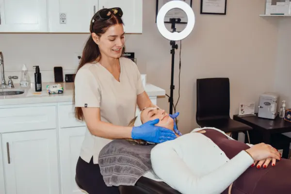Esthetician in beige scrubs wearing gloves treats a client lying on a treatment chair in a clinic, with a ring light in the background.