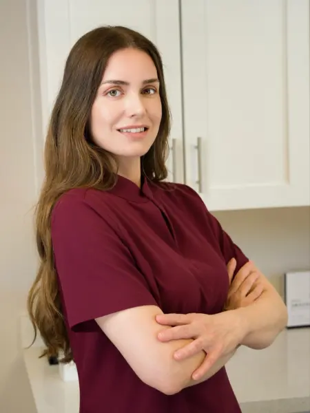 Professional woman with long brown hair wearing burgundy scrubs, arms crossed, standing in a bright kitchen.