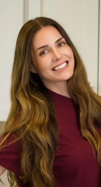 Smiling woman with long brown hair wearing a burgundy top, posing for a close-up selfie indoors.
